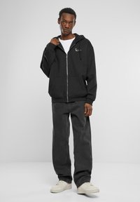Black zip-up hoodie with side pockets, featuring a white logo. Paired with loose-fitting dark jeans and white sneakers. Smooth fabric texture.