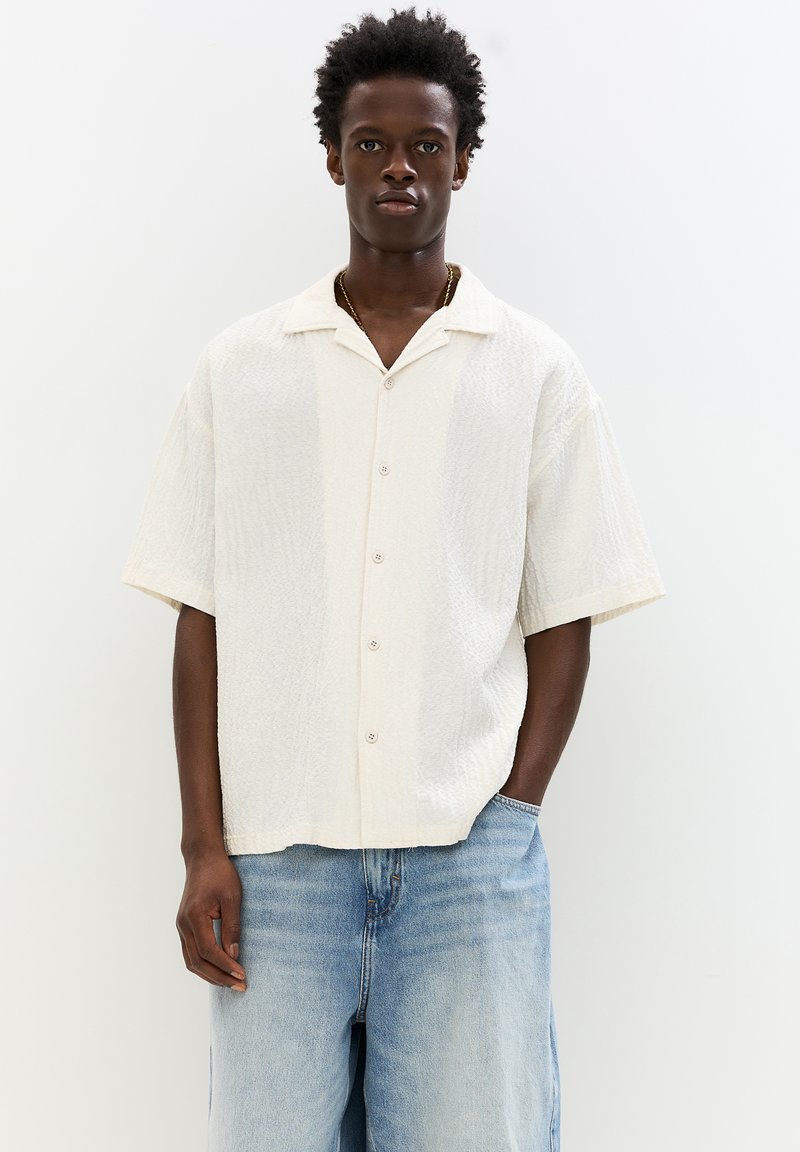 Short-sleeve white button-up shirt with a textured fabric, featuring a collar and an open, relaxed fit. Worn with light blue jeans.