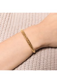 Złota skręcana bransoletka leży na nadgarstku w odcieniu skóry, z gładką fakturą na jasnobeżowym, dzianinowym tle.