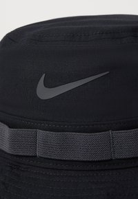 Chapeau seau noir en tissu avec une texture lisse. Présente un logo Nike gris et un band contrastant gris le long du bas.