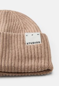 Beige ribbelgebreide beanie met opgerolde rand. Heeft een wit label met zwarte tekst "CPH STUDIOS." Zachte textuur en een snug fit.