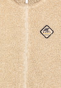 Beige fleecejacka med ett texturerat, lockigt material; har en framdragkedja och en Roxy-logotyp i diamantform.