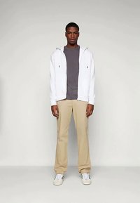 Vīrietis stāv uz viendabīgas fona, tērpies baltā ar rāvējslēdzēju, pelēkā T-kreklā, bēšos biksēs un baltās kedās ar melnām svītrām.