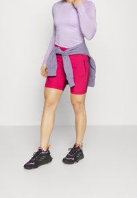 Vaaleanpunaiset shortsit, joissa on sivuvelcro-tasku, yhdistettynä vaaleanliilaan pitkähihaiseen puseroon ja mustiin urheilujalkineisiin, joissa on vaaleanpunaisia yksityiskohtia.