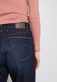 Jeans de mezclilla azul oscuro con un diseño distintivo en el bolsillo trasero y un parche de cuero marrón en la cintura visibles, combinados con una camiseta de manga larga rosa.