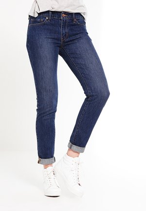 Skinny dark blue denim jeans with rolled cuffs, featuring a standard five-pocket design and subtle stitching details. Worn with white sneakers.