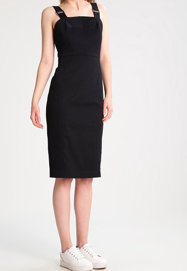 Kobieta ubrana w dopasowaną czarną sukienkę bez rękawów sięgającą do kolan, z klamrowymi ramiączkami i białymi trampkami, stojąca na tle jednolitą.