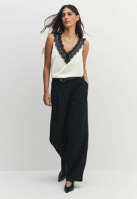 Balts satīna tops ar melnu mežģīņu apdari, kombinēts ar brīviem melniem biksēm un norādītām melnām kurpēm. Modelis stāv, rokas ieliktas kabatās.