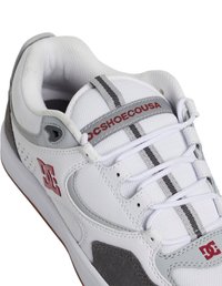 Bílé tenisky s šedými semišovými akcenty, texturovaným síťovaným svrškem, šněrováním a červeným brandingem DC Shoe Co. Obsahují černé pruhy na špičce.