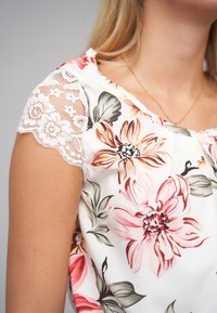 Bloes met bloemenpatroon en korte kanten mouwen, voorzien van grote roze en oranje bloemen op een witte achtergrond en een licht gerimpelde halslijn.