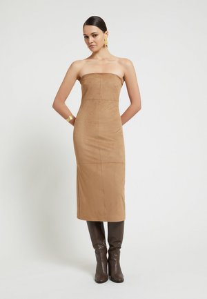 Donna con capelli scuri in un abito midi beige senza spalline e stivali marroni al ginocchio, con orecchini e bracciale d’oro, in piedi su sfondo bianco.