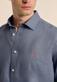 Camisa de botones azul claro con cuello, botones blancos y detalle de logo bordado rojo en el lado izquierdo del pecho. Fabricada en tejido texturizado.