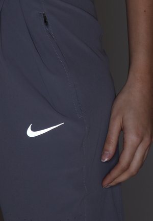 Zbliżenie na rękę spoczywającą obok ciemnych sportowych szortów z kieszenią na zamek i odblaskowym białym logo Nike swoosh.