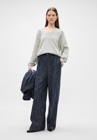 Sweater oversized cinza claro com decote em V, combinado com calças largas em padrão pied-de-poule azul escuro e um blazer a condizer, sapatos de salto pretos.