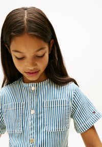Camicia a maniche corte blu e bianca a righe con scollatura tonda, dotata di due tasche frontali e chiusura a bottoni. Tessuto in cotone con una leggera trama.
