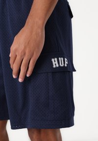Námornícky modré sieťované šortky s textúrovaným povrchom, so bočnou kapsou a vyšitými písmenami "HUF" v šedej farbe na klapke vrecka.