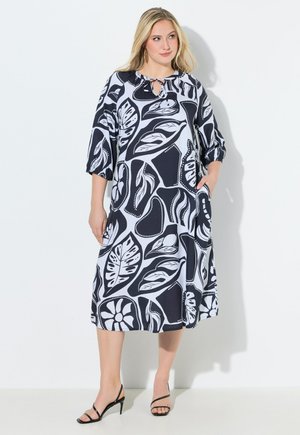 Kvinde iført en sort og hvid midikjole med bladmønster og trekvartærmer samt sorte sandaler med stropper, stående mod en hvid baggrund.