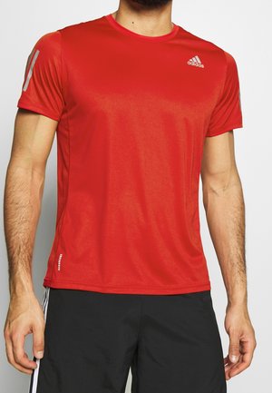 Mann trägt ein rotes Adidas-Sportshirt mit kurzen Ärmeln und schwarze Shorts mit weißen Streifen, steht vor hellem Hintergrund.