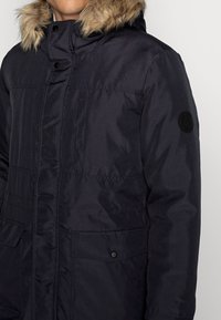 Marinblå pufferjacka med en huva kantad av fäll, knappförslutning fram, två stora fickor och en svart cirkulär logotyp på ärmen.