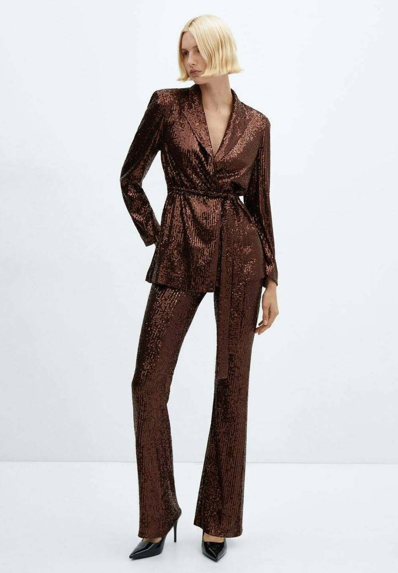 Donna che indossa un blazer scuro con paillettes e cinture abbinate, insieme a pantaloni coordinati e tacchi alti neri a punta, in posa davanti a uno sfondo chiaro e uniforme.