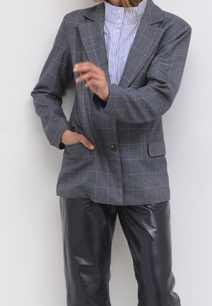 Szary, w kratkę marynarka z jednym guzikiem, klapami oraz dwoma przednimi kieszeniami, w połączeniu z błyszczącymi, czarnymi skórzanymi spodniami.