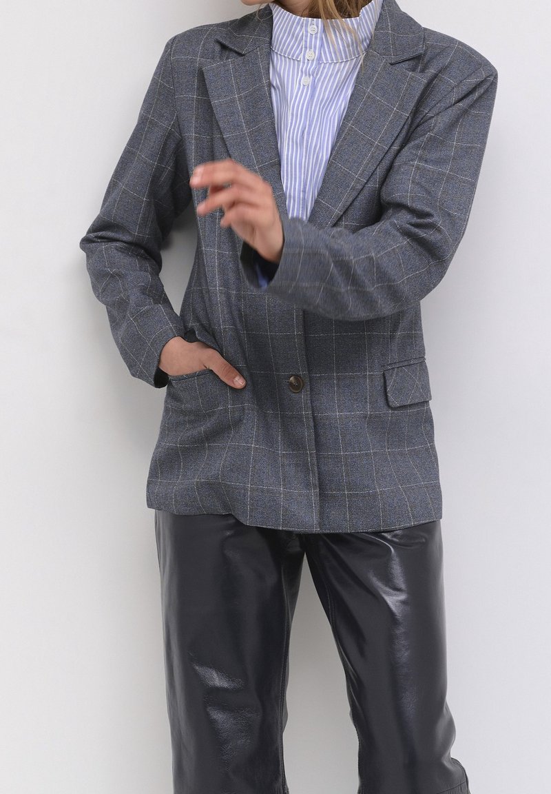 Szary, w kratkę marynarka z jednym guzikiem, klapami oraz dwoma przednimi kieszeniami, w połączeniu z błyszczącymi, czarnymi skórzanymi spodniami.
