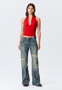 Raudonas halterio tipo viršus su giliais iškirpimais, derinamas su išblukusiais mėlynais plačiomis kojomis džinsais, turinčiais nudėvėtų vietų ir geltonų dažų purslų.