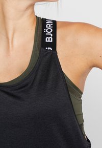 Zbliżenie osoby ubranej w warstwowe, zielone i czarne sportowe topy bez rękawów, z widocznym czarnym paskiem z białym napisem.