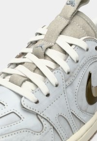 Gros plan sur une chaussure Nike beige clair et blanche avec des lacets blancs, des détails cousus, et le logo Nike métallique doré sur le côté.