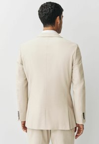 Beige op maat gemaakte blazer met notch-revers, een enkele split en zwarte knoopaccenten. Gladde stof met een gestructureerde pasvorm. Eenvoudige achteraanzicht.