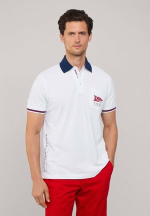 Witte polo met marineblauwe kraag, rode accenten en een zak op de borst. Heeft korte mouwen en verticale tekstdetails langs de zijkant.