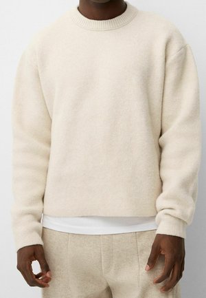 Béžový vlněný svetr s žebrovaným kulatým výstřihem, spadenými rameny a měkkým, texturovaným povrchem, doplněný o bílé spodní tričko.