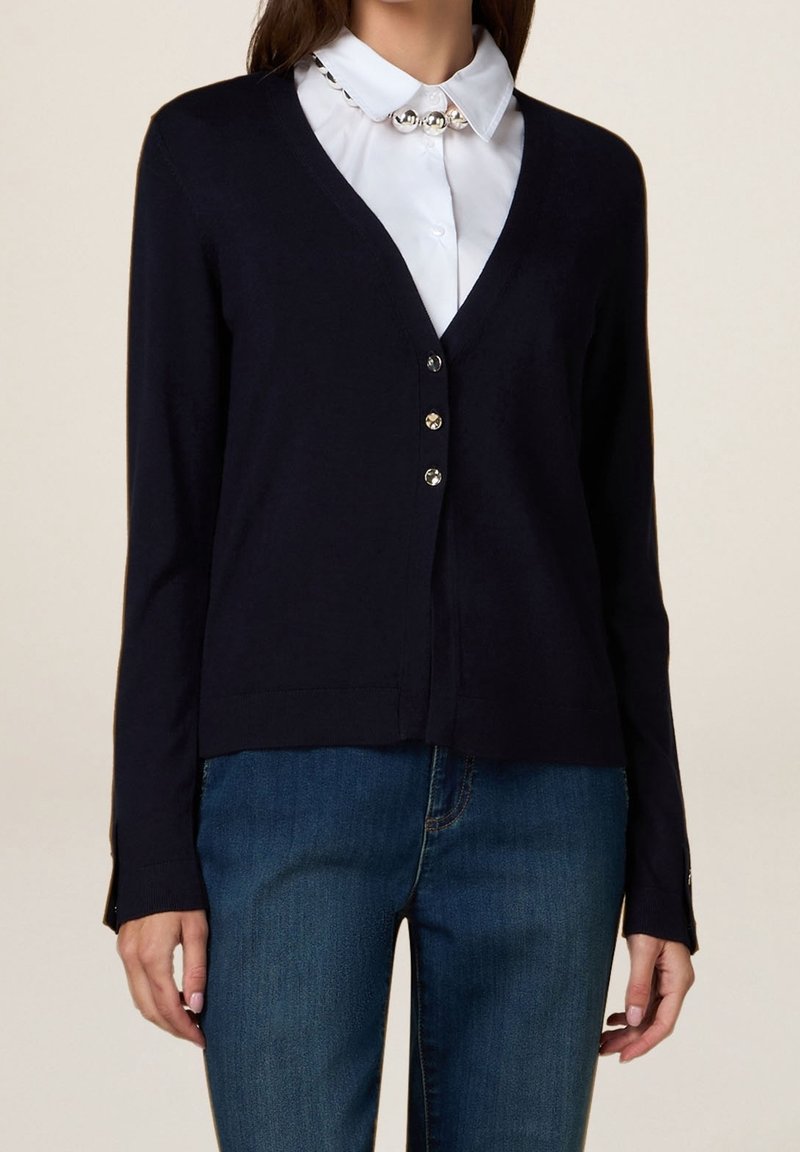 Femme portant un cardigan noir à col en V avec trois boutons sur une chemise blanche à col avec des ornements en perles, associé à un jean bleu.