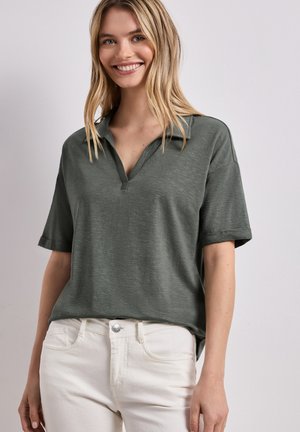 Jonge vrouw lacht, draagt een donker groene korte mouwen poloshirt en off-white broek, staand tegen een lichtgrijze achtergrond.