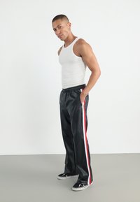 Pantaloni de trening negri, lucioși, cu dungi verticale roșii și albe, talie elastică și buzunare laterale. Modelul poartă un top alb și adidași.