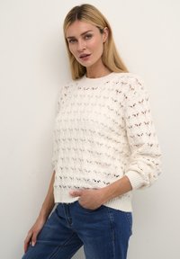 Maglione in crema lavorato a maglia con motivo traforato, scollo rotondo e polsini a costine. Abbinato a jeans blu, che mostrano una vestibilità comoda.