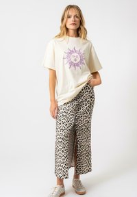 T-shirt couleur crème avec un graphisme de soleil violet, associé à une jupe léopard mi-longue avec une fente à l'avant et des baskets beiges.
