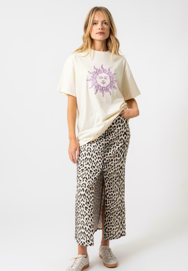 T-shirt couleur crème avec un graphisme de soleil violet, associé à une jupe léopard mi-longue avec une fente à l'avant et des baskets beiges.