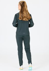 Donkergroene sportjack en bijpassende broek. Bevat ritsaccenten, reflecterende details en een zachte, rekbare stof voor beweging.