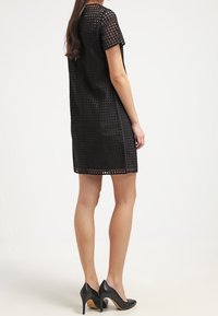 Vestido negro de manga corta con un diseño texturizado y perforado, de corte suelto, combinado con zapatos de tacón altos negros.