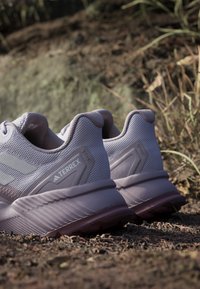 Paar hellgraue Adidas Terrex Trailrunning-Schuhe, die auf einem Kiesweg mit natürlichem Outdoor-Hintergrund liegen.