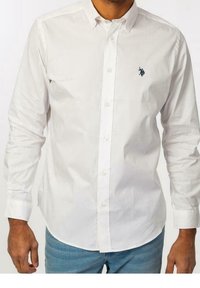 Camicia bianca a maniche lunghe con colletto a punta e logo ricamato blu scuro sul lato sinistro del petto. Jeans in denim chiaro visibili.