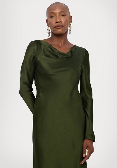 Langharige jurk in diep groene satijn met een gedrapeerde halslijn. Heeft een soepele textuur en een aansluitende silhouet. Statement gouden kettingoorbellen.