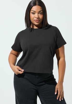 Mujer con cabello lacio y oscuro, vestida con una camiseta negra de manga corta y pantalones negros, de pie frente a un fondo claro.