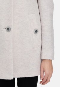 Helle graue Wolljacke mit weicher Textur, ausgestattet mit zwei vorderen Knöpfen und einer Seiten- Tasche, teilweise auf einer Hand vor einem schlichten Hintergrund gezeigt.