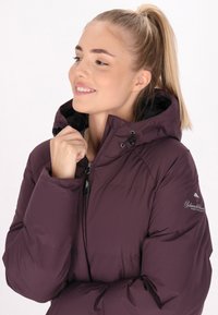 Giacca isolante viola con collo alto, cappuccio regolabile con cordino e fodera nera. Presenta una chiusura frontale con zip e ricamo del logo sulla spalla.