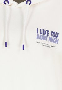 Witte hoodie met paarse trekkoordaccenten. Voorzijde met tekst: „IK MAG JE HEEL ERG” en „VRIENDSCHAP GAAT OVER KWALITEIT, NIET KWANTITEIT.”