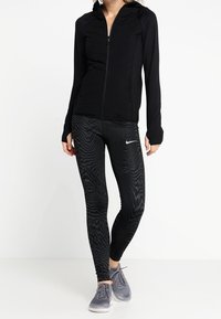 Schwarze Sportjacke mit Reißverschluss und glatter Textur, kombiniert mit schwarzen Leggings mit einem Wellenmuster, getragen mit grauen Sneakers.