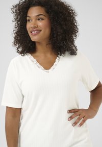 Haut blanc à manches courtes avec un design à côtes texturé, présentant une bordure en dentelle le long du décolleté en V pour une touche de détail supplémentaire.