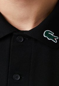 Polo en coton noir avec un tissu texturé, doté d'une patte de boutonnage à trois boutons et d'un logo crocodile vert brodé sur le col.